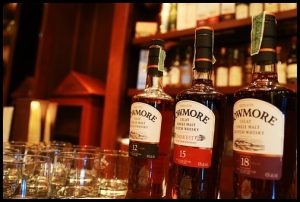 Three bottles of Bowmore whisky on a bar top with several whisky glasses beside them and blurred shelves filled whisky in the background.