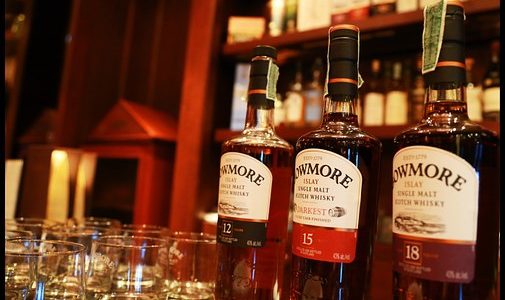 Three bottles of Bowmore whisky on a bar top with several whisky glasses beside them and blurred shelves filled whisky in the background.